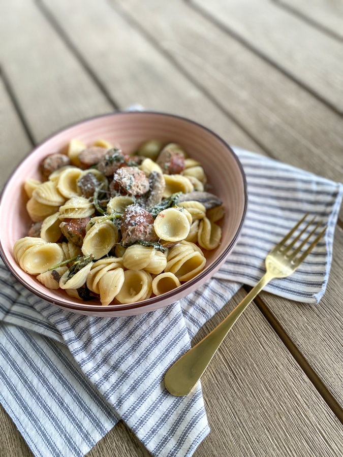 bowl of pasta with sausage and mushrooms over a gray napkin with a gold fork